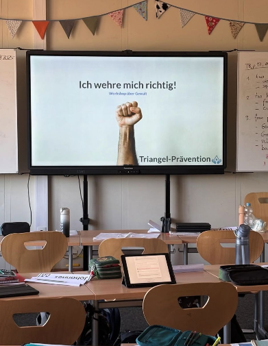 Klassenzimmer vor einem Triangel-Prävention-Workshop: Auf dem Smartboard erscheint die Folie „Ich wehre mich richtig! – Workshop über Gewalt“ mit einer erhobenen Faust.